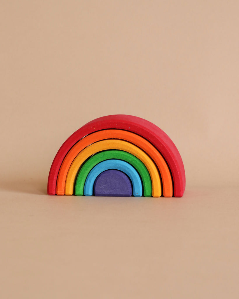 This Grimm's Small Wooden Rainbow, crafted from non-toxic wood, features six separate, colorful pieces arranged in a semi-circle. The colors—red, orange, yellow, green, blue, and purple—form a charming rainbow tunnel against a solid beige background. This versatile toy delights and engages.