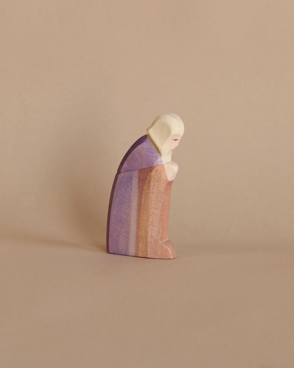 A minimalist Ostheimer Joseph wooden figurine of a person in prayer, featuring a hooded cloak painted in soft purple and peach hues against a plain beige background.