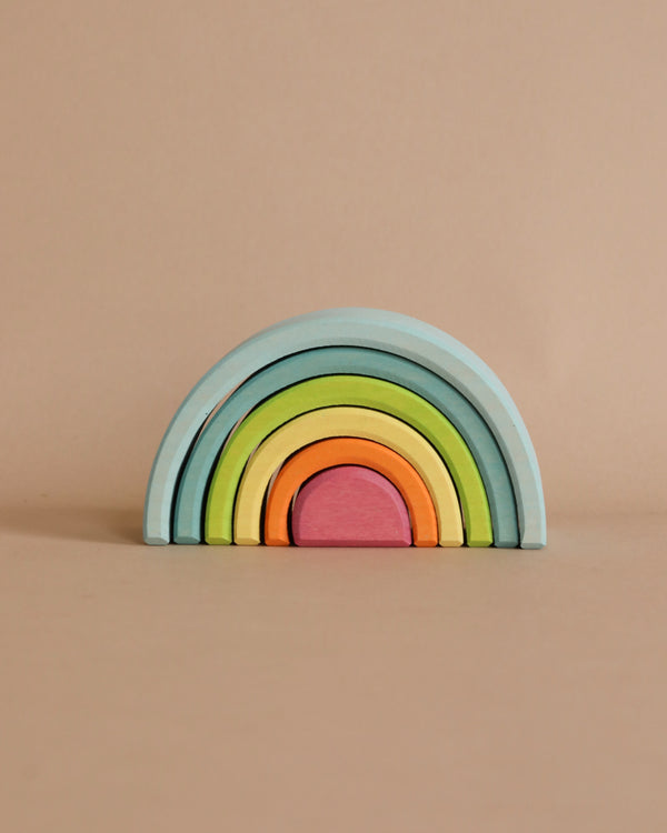 A Grimm's Small Wooden Rainbow, Pastel with six pastel arches. The largest arch is light blue, followed by green, yellow, orange, pink, and a small red arch at the center. This versatile toy is set against a beige background with no other objects in the scene.