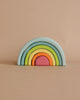 A Grimm's Small Wooden Rainbow, Pastel with six pastel arches. The largest arch is light blue, followed by green, yellow, orange, pink, and a small red arch at the center. This versatile toy is set against a beige background with no other objects in the scene.