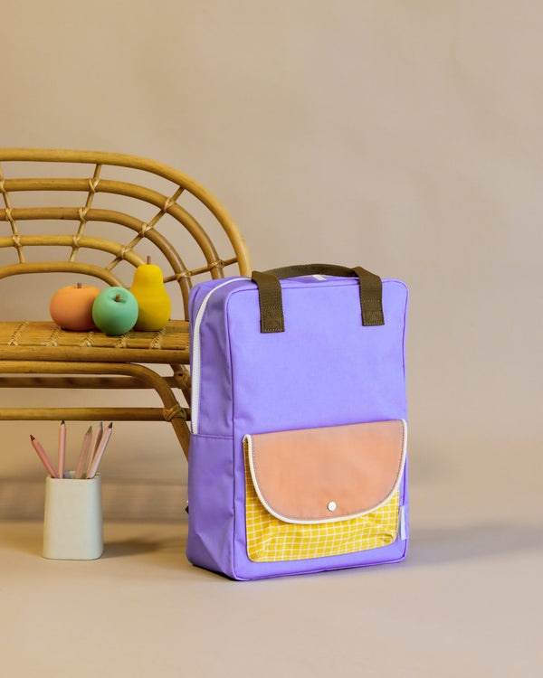 A purple Sticky Lemon Backpack Large with a yellow front pocket, and a tan flap made of recycled RPET stands upright beside a bamboo chair and a container holding art supplies.