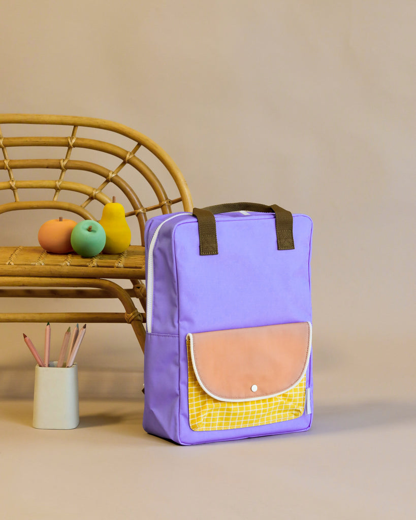 A purple Sticky Lemon Backpack Large with a yellow front pocket, and a tan flap made of recycled RPET stands upright beside a bamboo chair and a container holding art supplies.