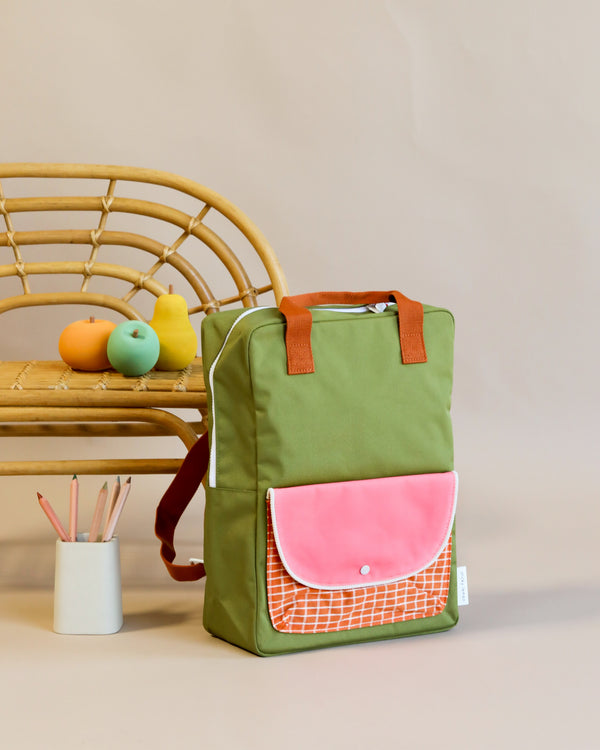 A Sticky Lemon backpack large with a coral flap pocket made of waterproof nylon rests against a woven chair. On the chair, there are colorful toy blocks and a white cup holding pencils. A neutral background enhances the scene.