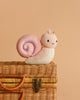 The Cuddle + Kind Baby Snail (Peony) features a plush snail with a pink spiral shell and a tiny crown, perched on a woven wicker basket against a beige background—an heirloom-quality doll ideal for gifting.