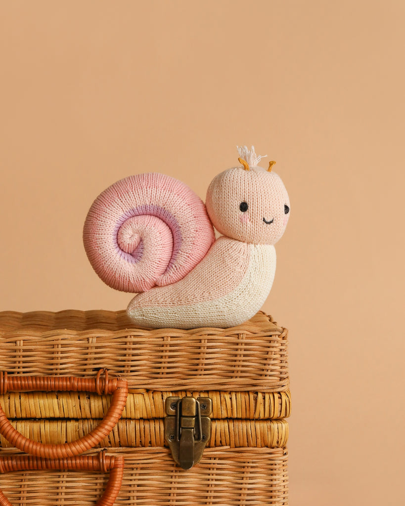 The Cuddle + Kind Baby Snail (Peony) features a plush snail with a pink spiral shell and a tiny crown, perched on a woven wicker basket against a beige background—an heirloom-quality doll ideal for gifting.
