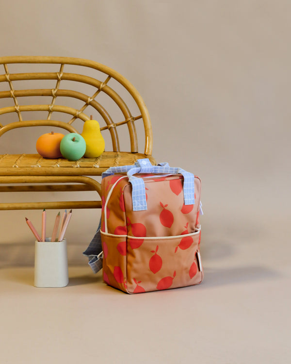 A Sticky Lemon Backpack Small | Farmhouse | Special Edition with a peach print design, featuring a durable YKK zipper, stands in front of a rattan chair. On the chair, there are two artificial fruits, and a white container holds