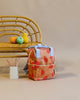 A Sticky Lemon Backpack Small | Farmhouse | Special Edition with a peach print design, featuring a durable YKK zipper, stands in front of a rattan chair. On the chair, there are two artificial fruits, and a white container holds