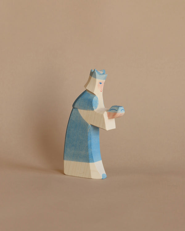 A handcrafted Ostheimer Blue King With Crown wooden figurine painted to resemble a person wearing a blue and white outfit, depicted in a praying or contemplative pose, against a plain beige background.