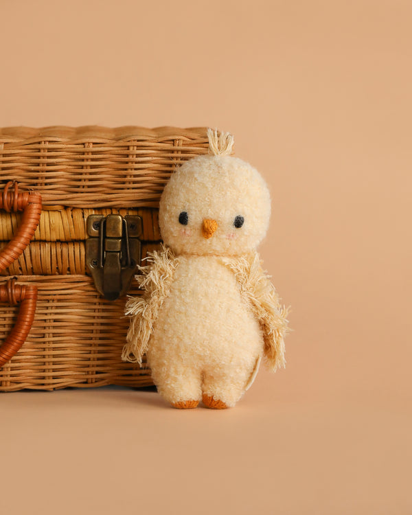 The Cuddle + Kind Baby Bouclé Chick (Buttercup), a small plush toy with a fuzzy feel, stands next to a wicker basket on a beige background—ideal for collectors of adorable hand-knit baby animals and collectible plush.