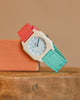 The Mini Kyomo Watch - Bloom, featuring a white octagonal face with pink and teal bands, is displayed on a peach block atop a wooden surface against a neutral background.