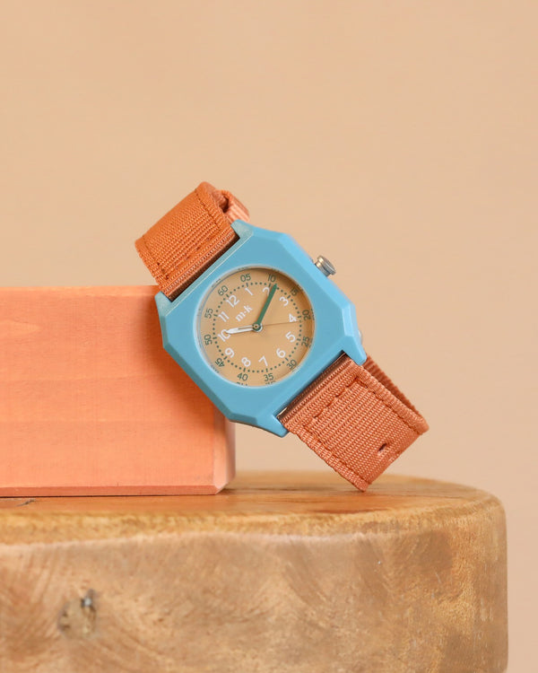 The Mini Kyomo Watch - Havana Sky, with a blue face and orange strap, rests on a peach block atop a wooden surface against a beige background.