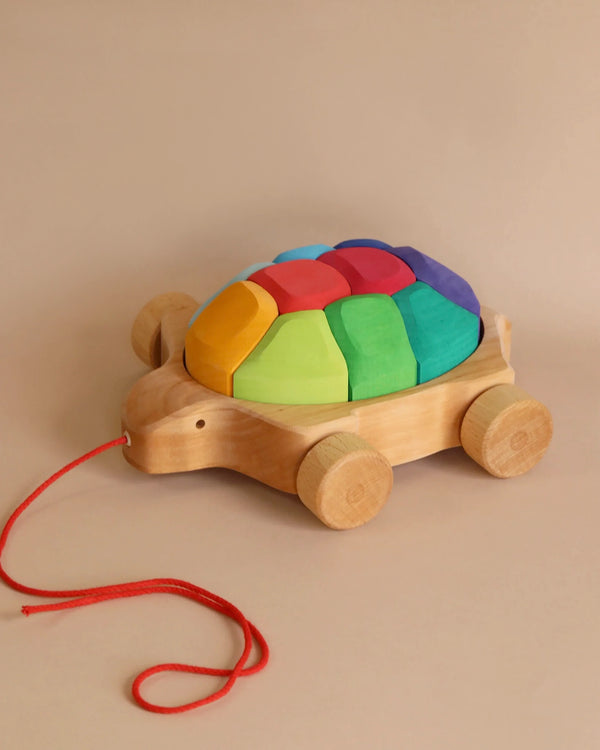 A Grimm's Rainbow Turtle Pull-Toy and Building Blocks shaped like a turtle with large wheels. The turtle's shell consists of colorful, interlocking, geometric pieces in red, yellow, green, blue, and purple—resembling a rainbow-colored tortoiseshell. A red string attached to the front helps develop fine motor skills as it is pulled along.