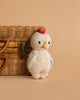 The Cuddle + Kind Baby Chicken, a small hand-knit chick with a red comb and yellow beak, stands in front of a closed wicker basket on a beige background.