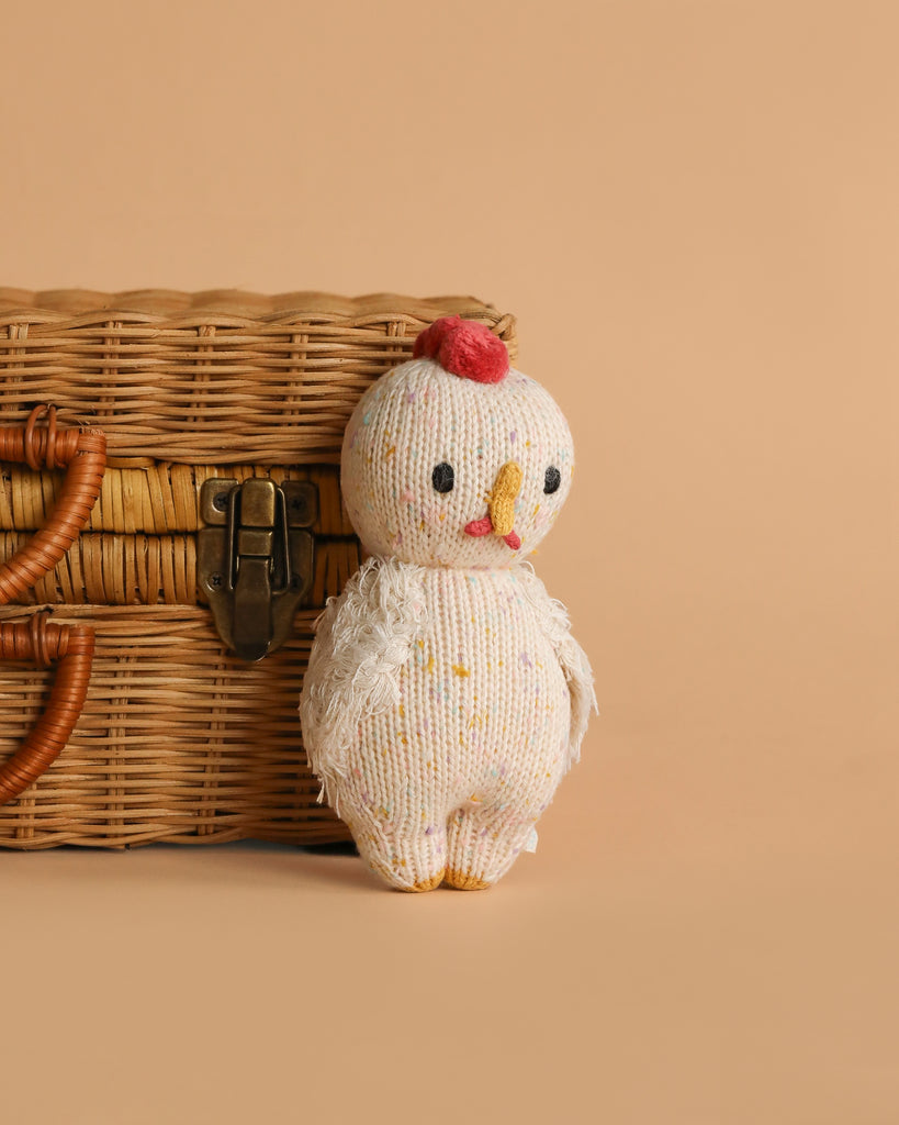 The Cuddle + Kind Baby Chicken, a small hand-knit chick with a red comb and yellow beak, stands in front of a closed wicker basket on a beige background.