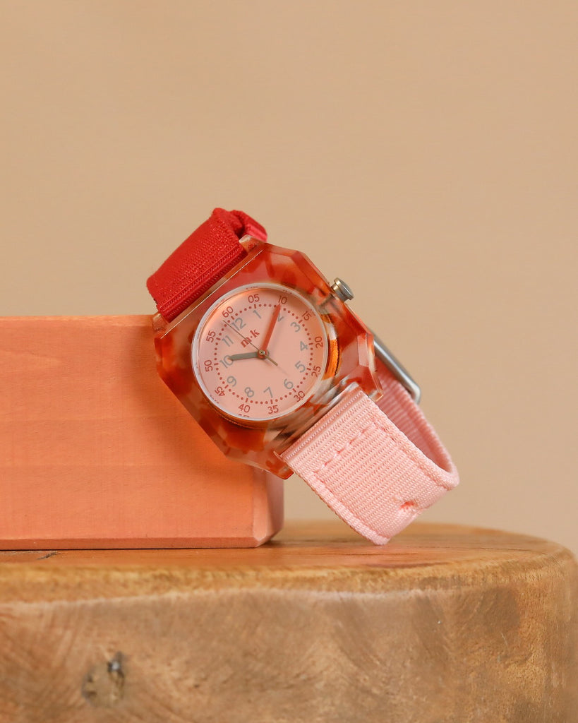 A Mini Kyomo Watch - Cinnamon Roll with a clear red plastic case sits on a salmon-colored block, displayed on a wooden surface against a beige background.