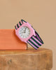 A Mini Kyomo Watch - Lavender Field with a white face and a pink, black, and green striped fabric band sits on an orange block on a wooden surface. It features precise Japanese movement.