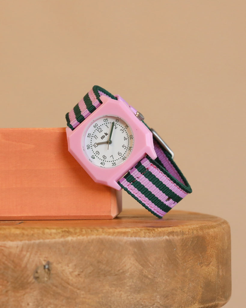 A Mini Kyomo Watch - Lavender Field with a white face and a pink, black, and green striped fabric band sits on an orange block on a wooden surface. It features precise Japanese movement.