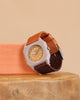 A Mini Kyomo Watch - Plum Cake with a hexagonal face, orange dial, and brown and orange fabric strap sits on a round wooden surface next to a rectangular orange block.