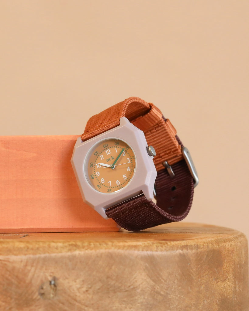 A Mini Kyomo Watch - Plum Cake with a hexagonal face, orange dial, and brown and orange fabric strap sits on a round wooden surface next to a rectangular orange block.