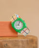 The Mini Kyomo Watch x Taxi Brousse - Color Notebook, with a green octagonal case and colorful plaid strap, sits on an orange block atop wood in a playful vintage style against a beige background.