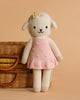Cuddle + Kind Eleanor the Lamb is a hand-knit doll, wearing a flower headband and pink dress with yellow dots, pictured before a wicker basket on beige. Lovingly handmade in Peru.