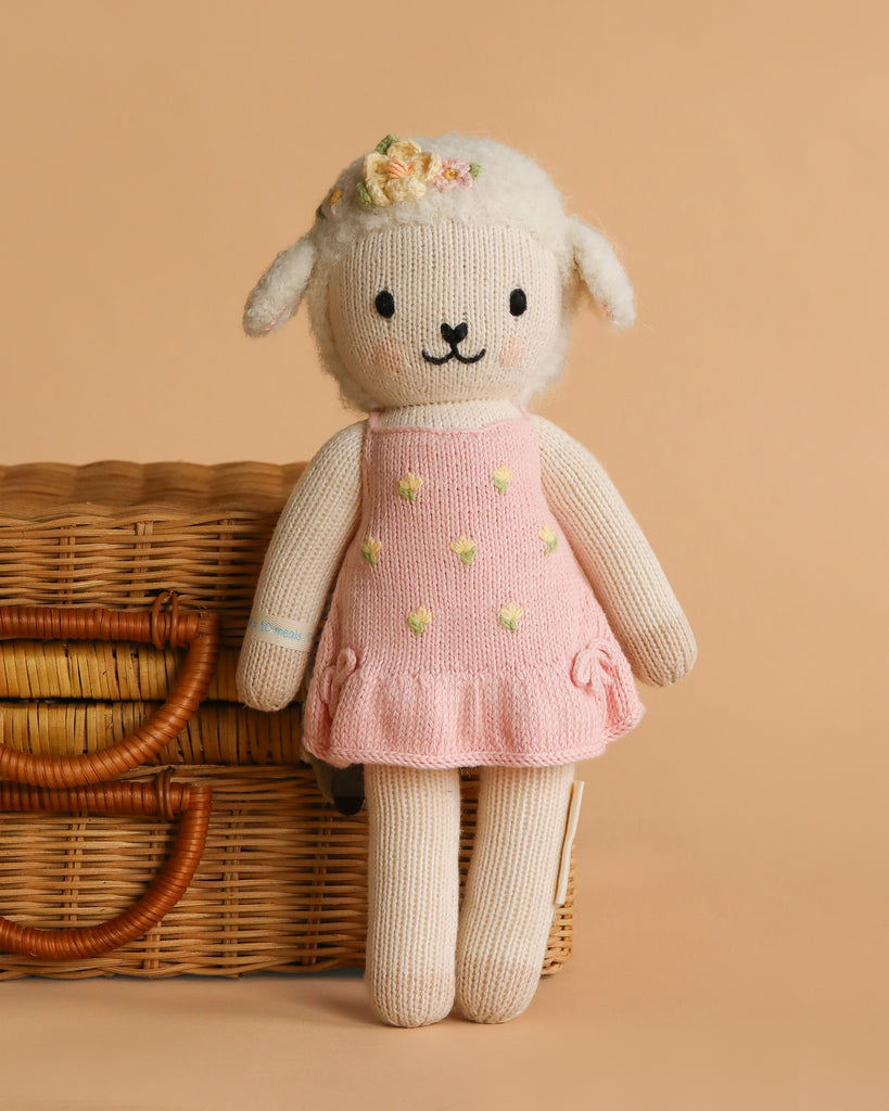 Cuddle + Kind Eleanor the Lamb is a hand-knit doll, wearing a flower headband and pink dress with yellow dots, pictured before a wicker basket on beige. Lovingly handmade in Peru.