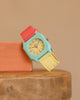 The Mini Kyomo Watch - Bubble Gum features a turquoise face, red and yellow straps, and a pink dial, displayed on a peach block atop wood with a beige backdrop.
