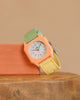 The Mini Kyomo Watch - Watercolor, featuring a pastel peach hexagonal face, green and yellow strap, white dial, and red numerals, is displayed on wood with a peach block in the background.
