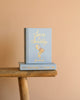 A light blue hardcover "Sense and Sensibility | Collector's Edition" with gold lettering and two hats is displayed on a rustic wooden bench against a peach background.
