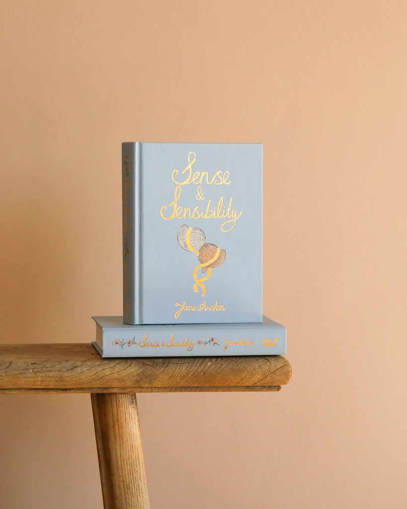 A light blue hardcover "Sense and Sensibility | Collector's Edition" with gold lettering and two hats is displayed on a rustic wooden bench against a peach background.