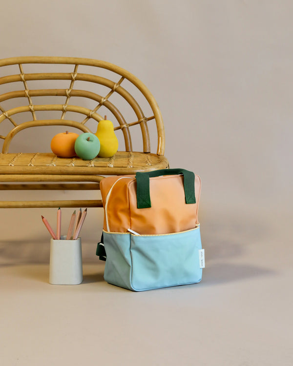 Sentence with product name: A Sticky Lemon Backpack Small in Meet Me In The Meadows color blocking pattern stands in front of a rattan bench with colorful faux fruits and a white pencil holder containing pink pencils, against a neutral background.