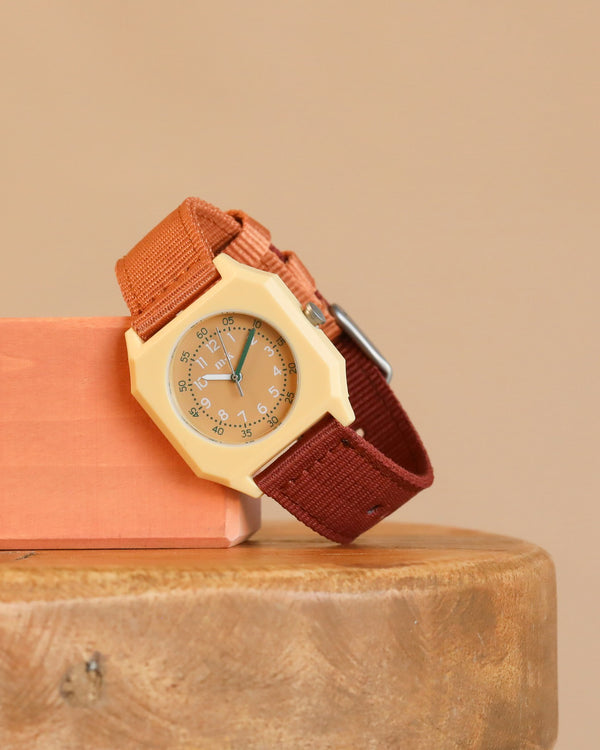 A Mini Kyomo Watch - Cherry Bomb with a beige octagonal face and brown fabric strap rests on wood, leaning against a peach block with a neutral beige background.