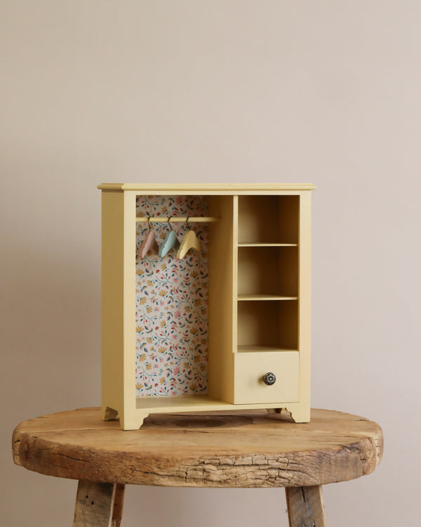 A small, yellow wooden Maileg Wardrobe, Large with an open door, displaying colorful floral interior lining and a shelf with neatly folded pastel-colored fabrics, placed on a rustic wooden table against a plain beige