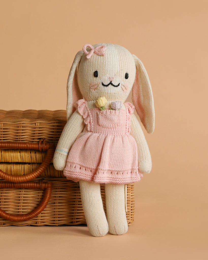 Cuddle + Kind Clover the Bunny (Blossom), a hand-knit doll in a pink dress and bow, stands before a wicker basket against a beige background.