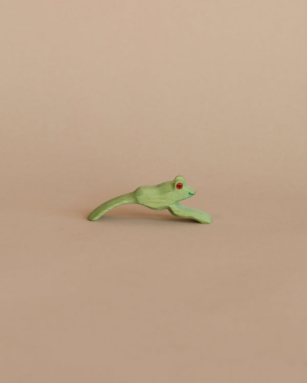 A small, green handcrafted Ostheimer Jumping Frog with red eyes, depicted mid-leap on a plain beige background.