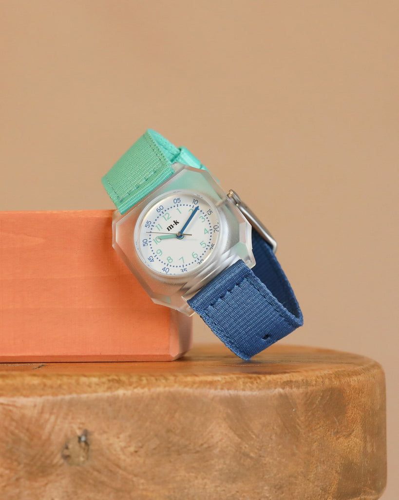 A Mini Kyomo Watch - Blue Ocean with a white face, blue numbers and hands, and a blue-mint green strap sits on an orange block on wood, against a beige background.