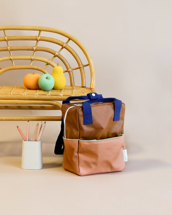 A stylish Sticky Lemon Backpack Small in Treehouse Brown with a YKK zipper stands upright next to a rattan bench with colorful faux fruit on top and a white cup holding pencils beside it, against a neutral beige background.