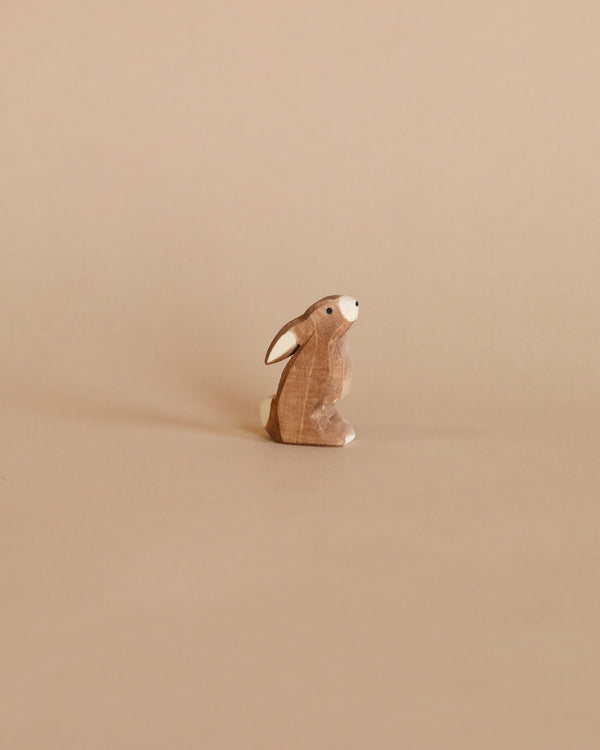 A small wooden Ostheimer Rabbit - Ears Low figurine stands isolated against a plain, light brown background. The rabbit is slightly textured, adding a rustic charm to its appearance.