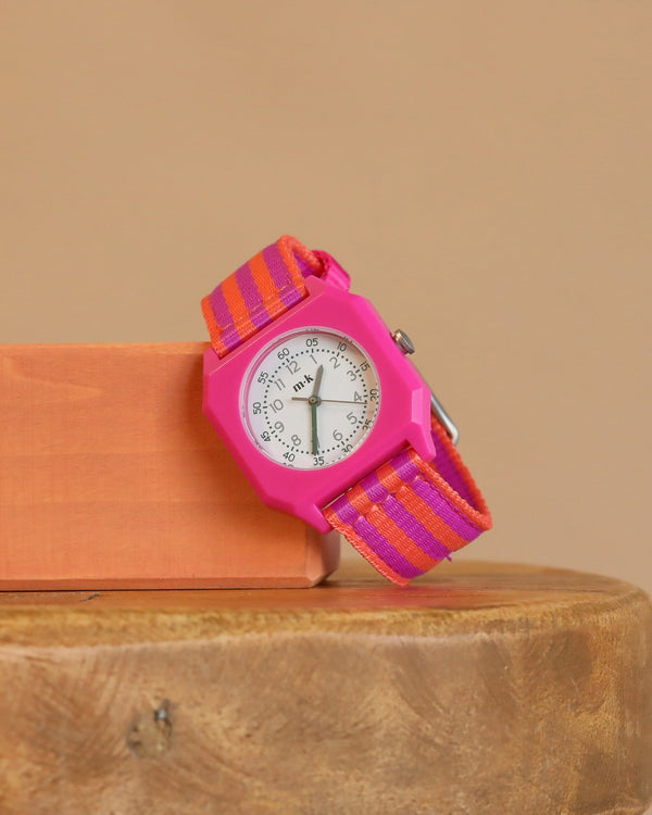 The Mini Kyomo Watch - Ruby Papaya, featuring Japanese movement and a pink face with a pink-orange striped fabric band, is displayed on an orange block against a neutral background and wooden surface.