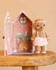 A hand-knit Cuddle + Kind Tiny Gingerbread - Sugarplum doll wearing a pink skirt and headband stands next to an open cardboard gingerbread house.