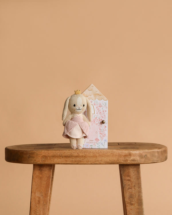 Cuddle + Kind Tiny Rose the Princess is a hand-knit plush bunny in a pink dress and crown, standing on a wooden bench before a pastel gift box—all handmade in Peru with fair trade practices, set against a plain beige background.