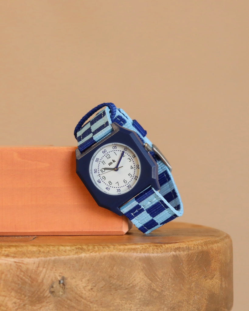 Mini Kyomo Watch - Blue Sunday - Blue Checkered stands upright on wood, leaning on an orange block with a beige background; its blue and white checkered band and blue bezel create a playful vibe for kids.