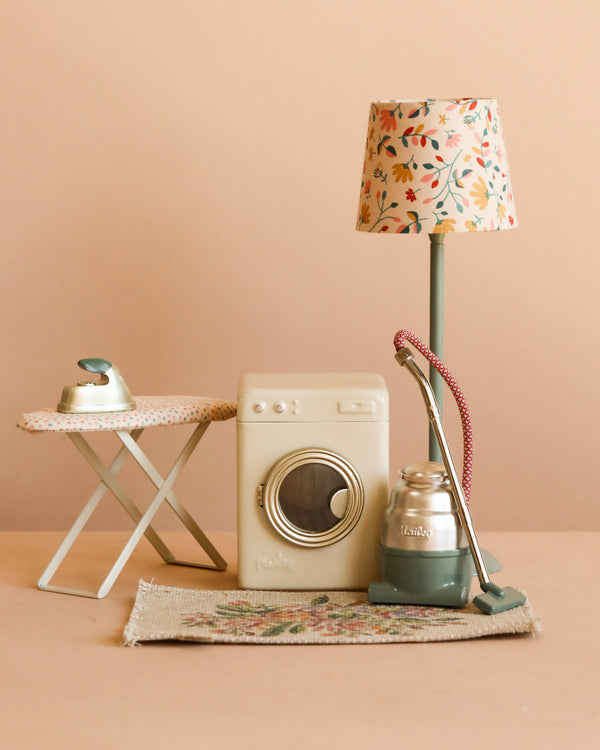 The Maileg Miniature Appliance Set features a washing machine, ironing board with iron, vacuum cleaner, tall lamp with floral shade, and small rug, all displayed against a beige background.