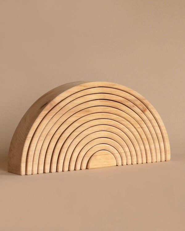 A Grimm's Large Natural Rainbow Stacker, crafted from non-toxic alder-wood, is composed of smoothly curved, nested arches graduated in size, forming a semi-circle. This natural tunnel's light brown color blends seamlessly with the beige background.