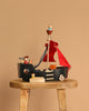 The Pirate Ship Set, featuring red sails and small figurines, sits on a rustic wooden stool against a plain beige background, making it an enchanting toy for children.