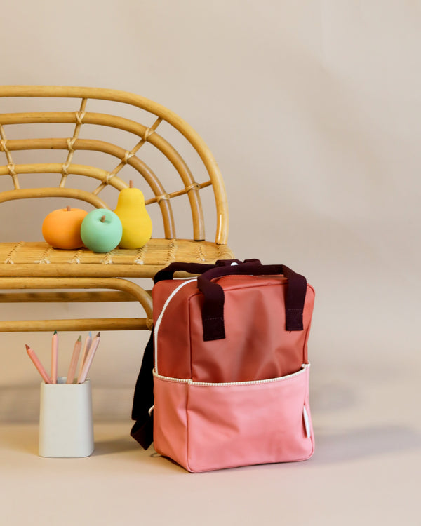 A Sticky Lemon Backpack Small with a YKK zipper stands on the floor next to a wicker bench, which holds a bowl with colorful fruit and a white cup filled with pencils. The background is plain and light.