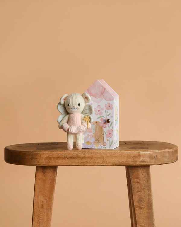Cuddle + Kind Tiny Lily the Fairy, a hand-knit doll in a pink dress, stands next to a pastel house-shaped box with floral designs on a wooden bench against a plain beige background.