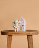 Cuddle + Kind Tiny Lily the Fairy, a hand-knit doll in a pink dress, stands next to a pastel house-shaped box with floral designs on a wooden bench against a plain beige background.