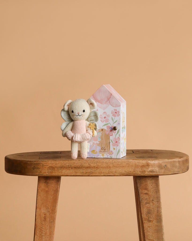Cuddle + Kind Tiny Lily the Fairy, a hand-knit doll in a pink dress, stands next to a pastel house-shaped box with floral designs on a wooden bench against a plain beige background.