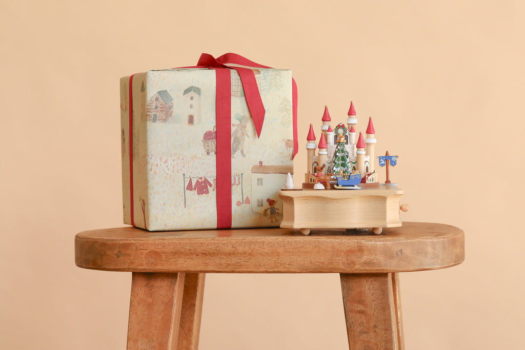 A wrapped gift tied with a red ribbon sits on a wooden stool next to a Christmas Castle Music Box featuring a castle, a Christmas tree, and small figures.